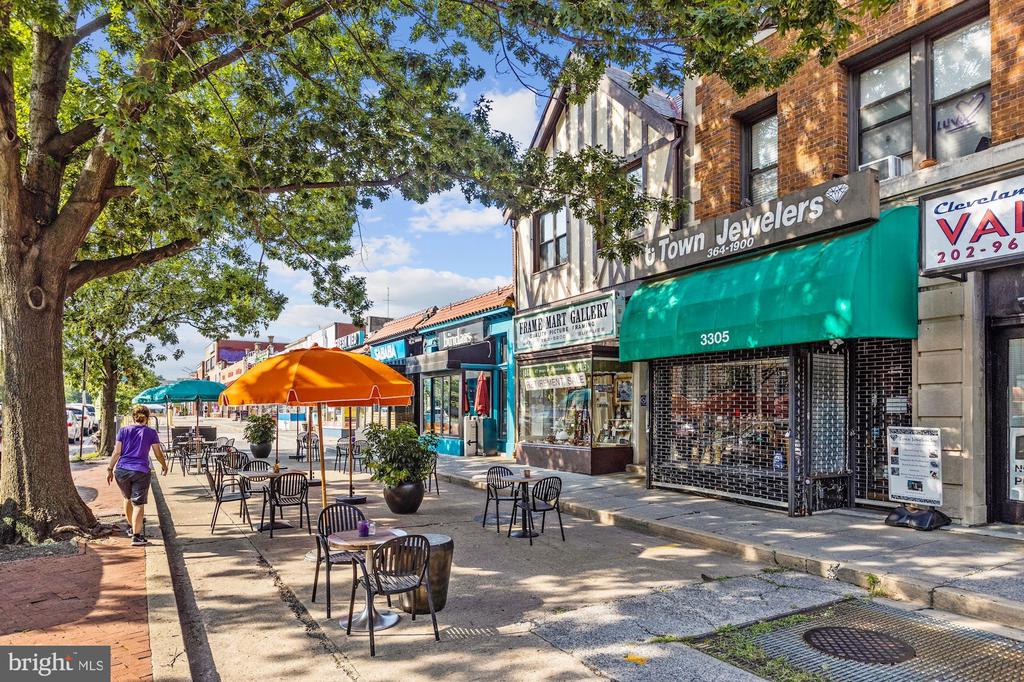 Tree-shaded commercial sidewalk with brick storefronts and outdoor café seating.