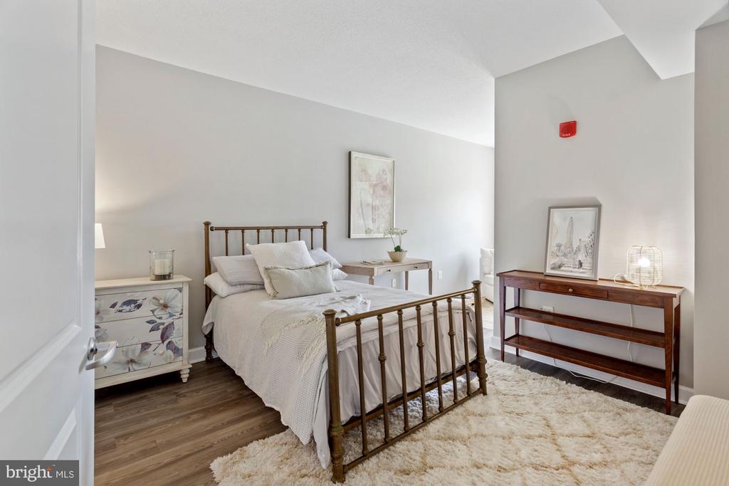 Staged bedroom with white bedding, a dark metal bed, floral bedside chest, and pale rug.