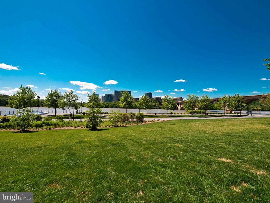 Riverside park with broad green lawn, river, bridge, and distant city skyline.
