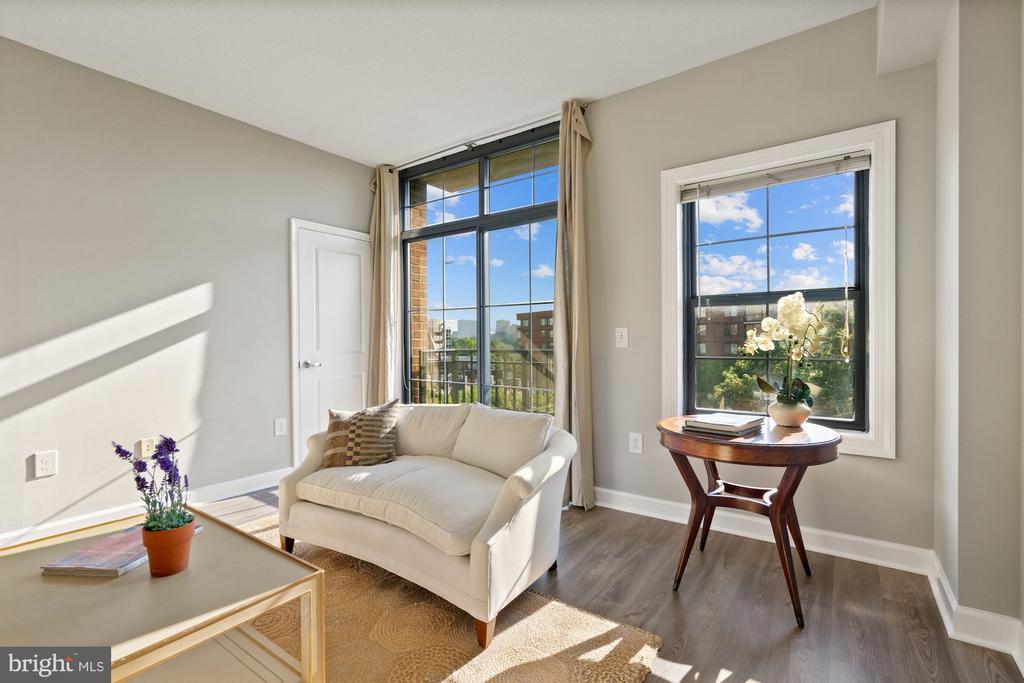 Bright staged living room with a white loveseat, tall windows, and views of nearby buildings.