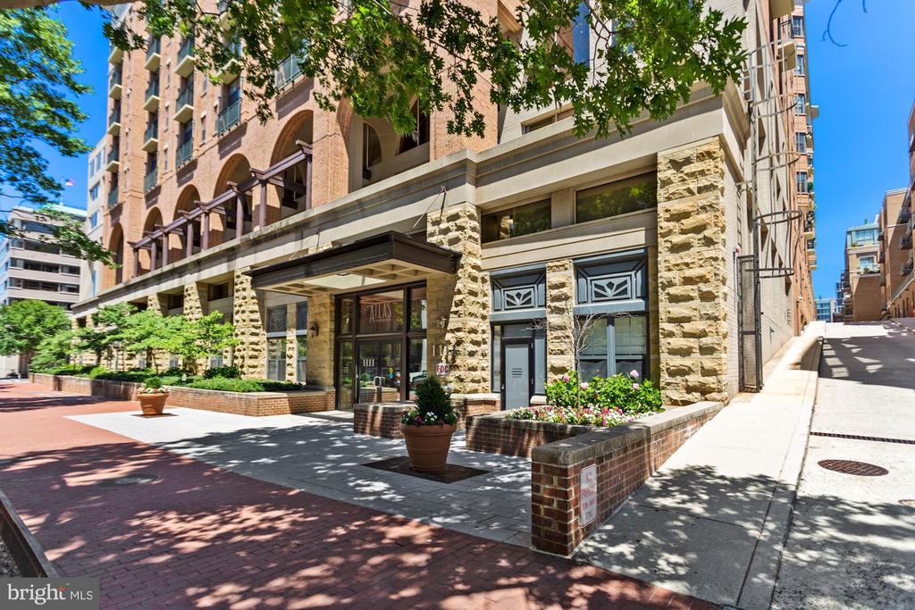 Residential building entrance with a dark canopy, stone facade, and flower-filled planters.