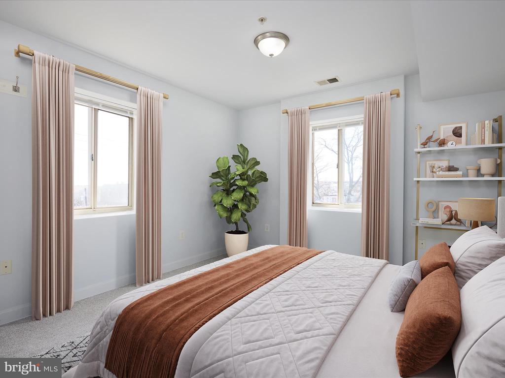 Staged bedroom with a white quilted bed, rust-colored accents, two curtained windows, and a potted plant.