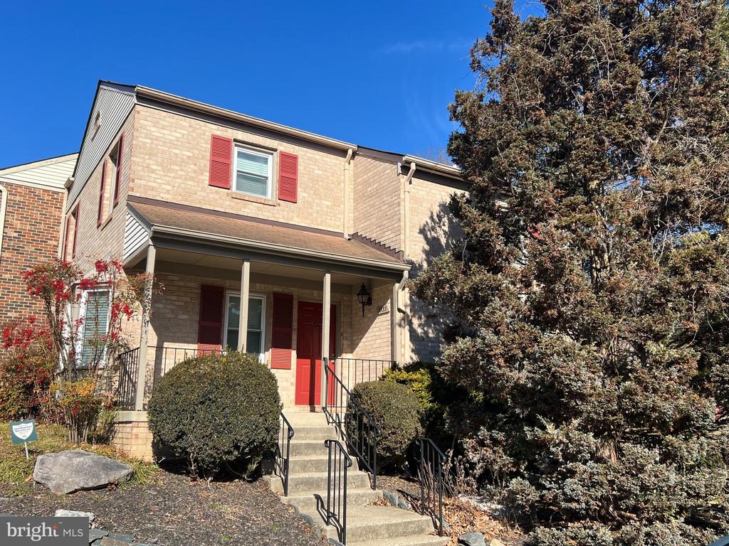 Two-story brick home with red shutters, a red front door, covered porch, and large evergreen tree.