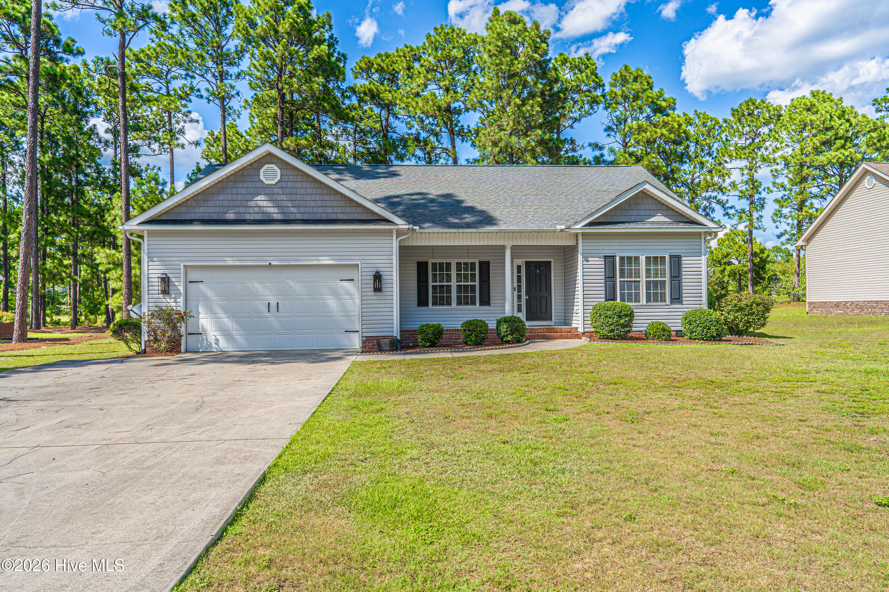 Gray single-story house with a two-car garage, brick accents, and tall pines behind it.