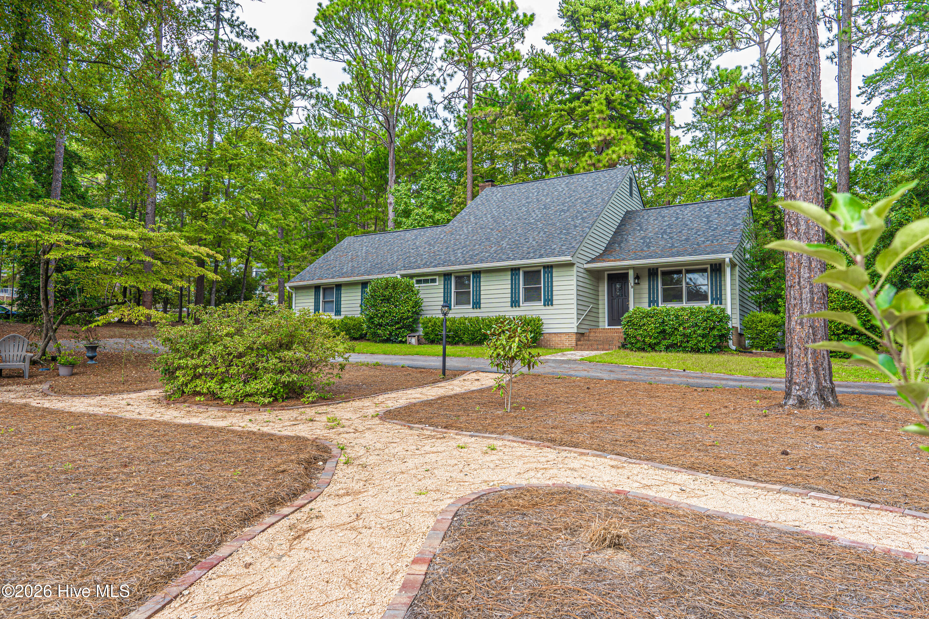 Single-story house among pine trees, with blue-green shutters and a curved gravel walkway.