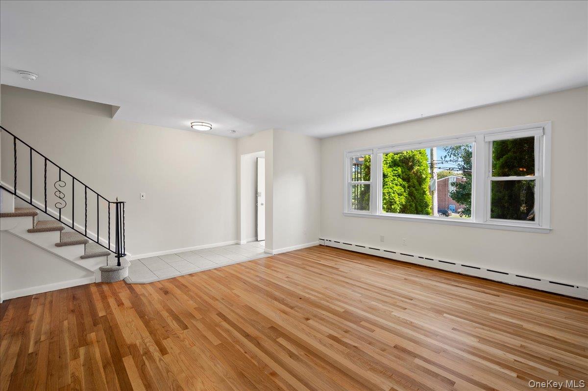 Empty living room with hardwood floors, a black-railed staircase, and a wide window overlooking greenery.