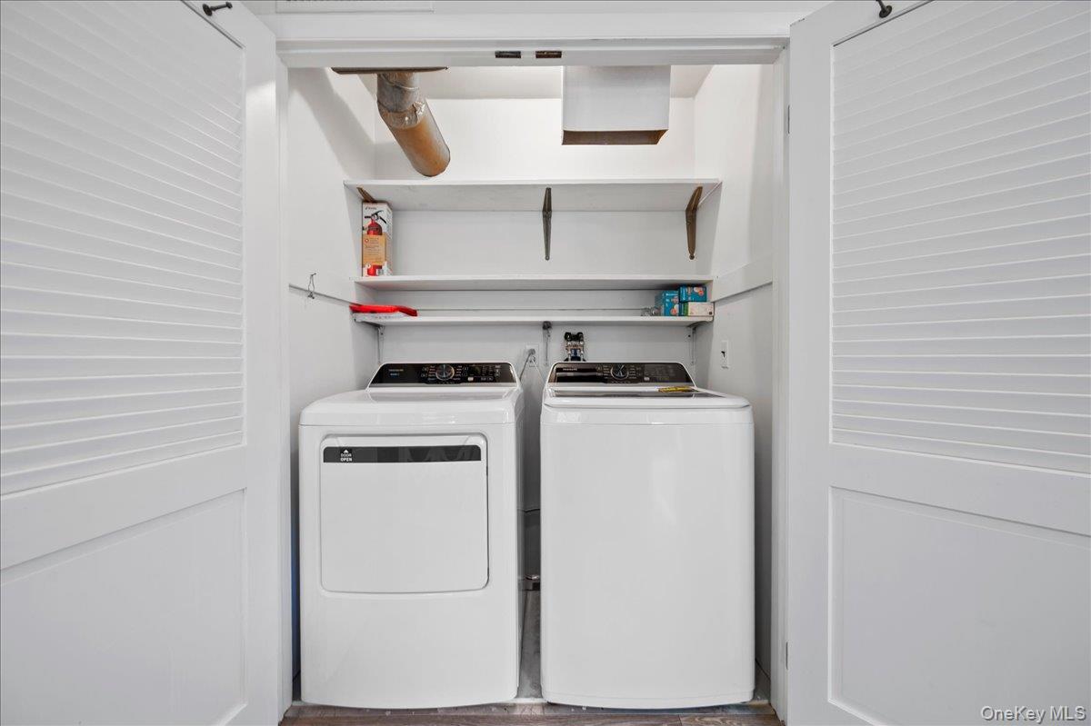 Laundry closet with a white dryer and washer beneath open shelves, behind louvered doors.