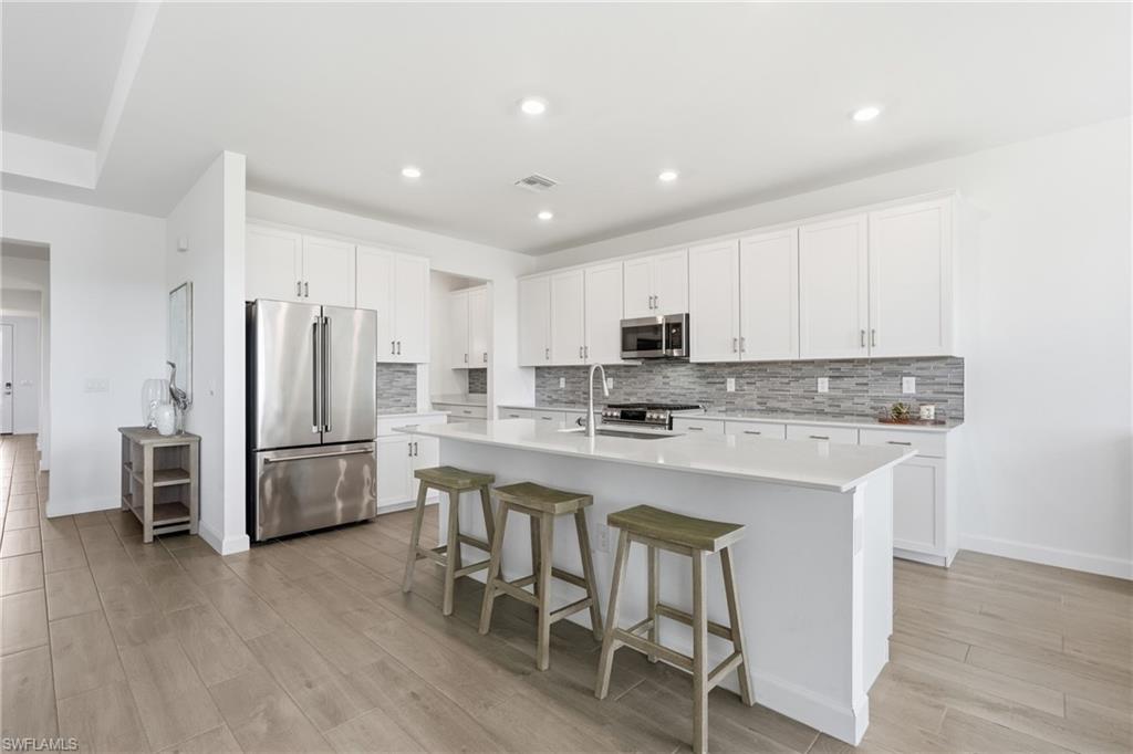Bright kitchen with a large white island, bar stools, stainless appliances, and gray tile backsplash.