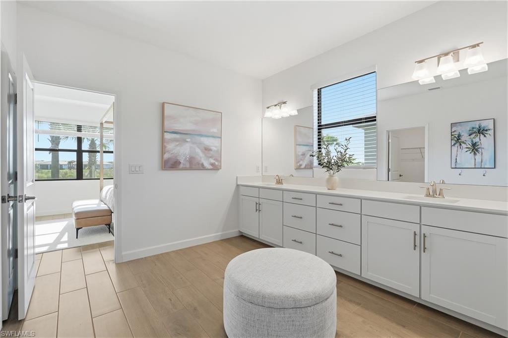 Bright bathroom with a long double vanity and a round upholstered stool beside an open bedroom doorway.