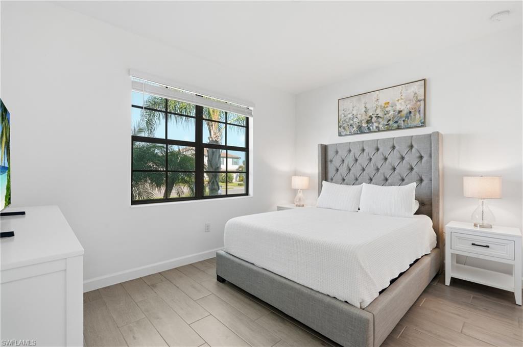 Staged bedroom with a gray upholstered bed, white bedding, and a window overlooking palm trees.