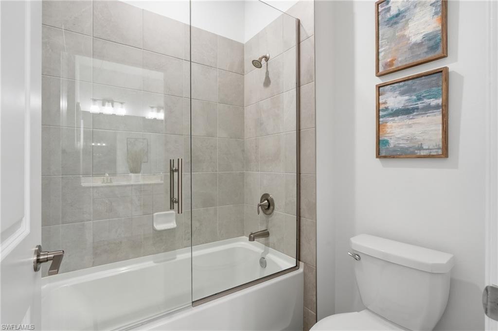 Bathroom with a glass-enclosed tub, gray tile walls, and two framed artworks above the toilet.