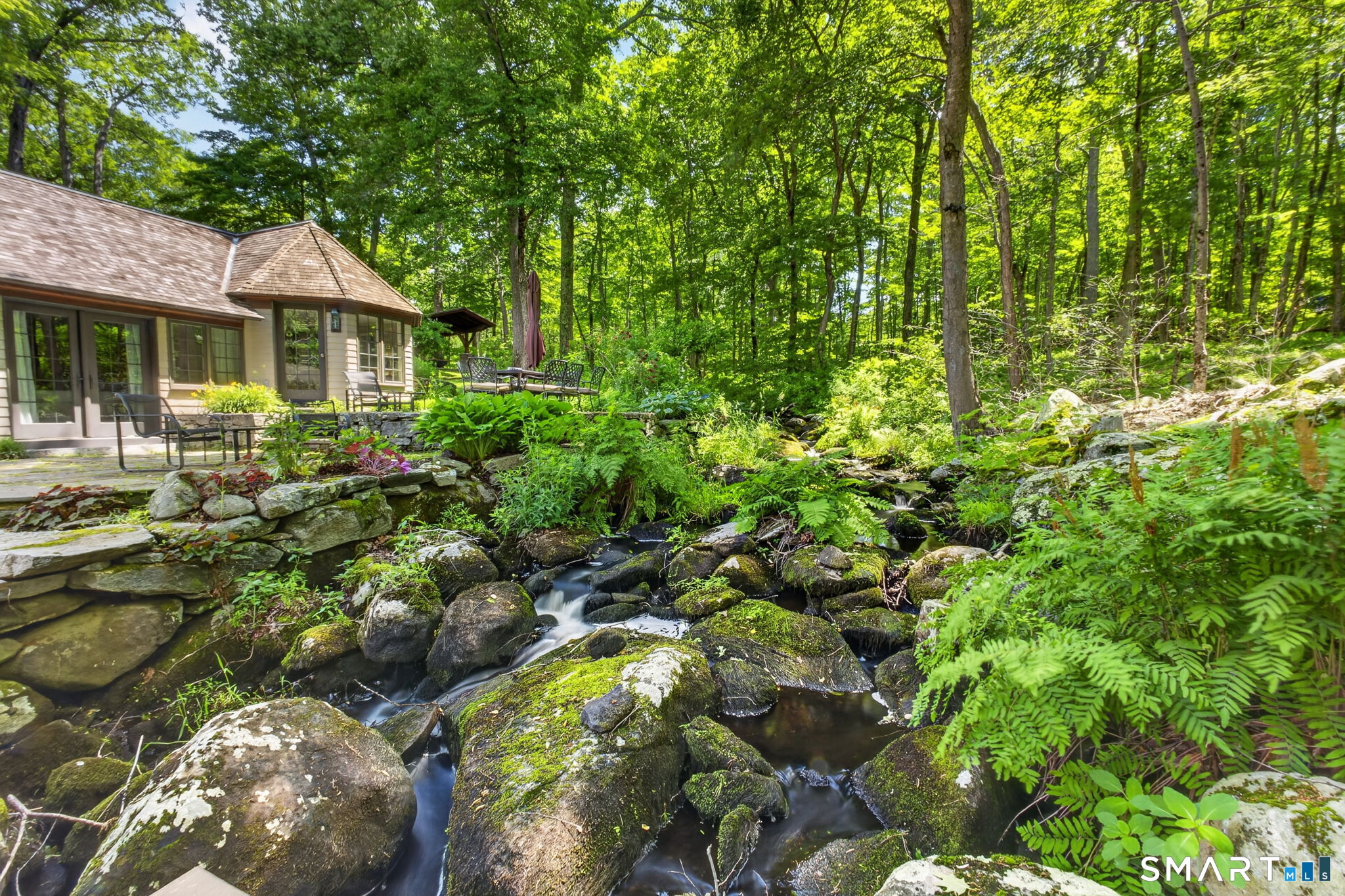 Wooded home patio beside a rocky stream, with stone walls and outdoor seating.