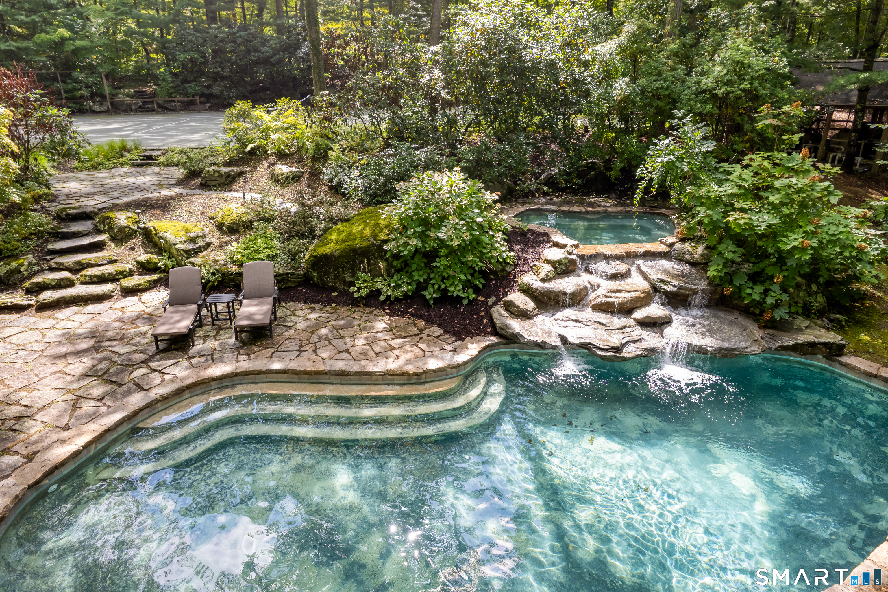 Swimming pool with a stone waterfall, surrounded by lush landscaping and a patio with chaise lounges.