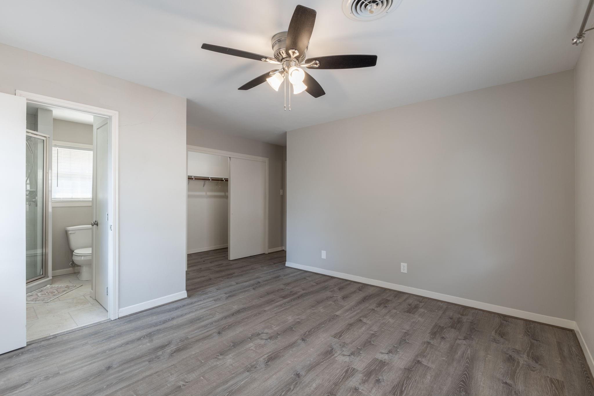 Empty bedroom with gray wood-look flooring, ceiling fan, open closet, and adjoining bathroom.