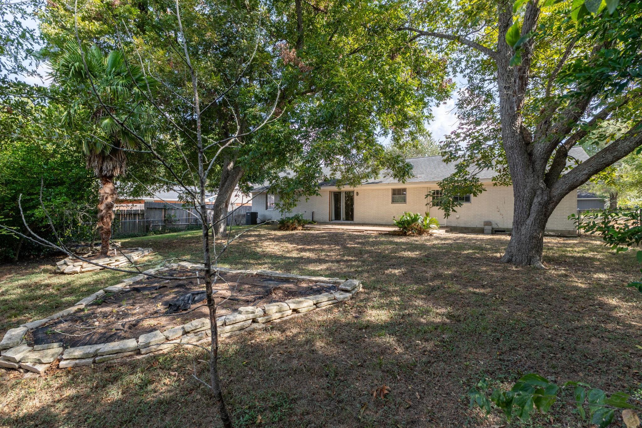 Single-story brick home behind a shaded backyard with mature trees and a stone-edged garden bed.