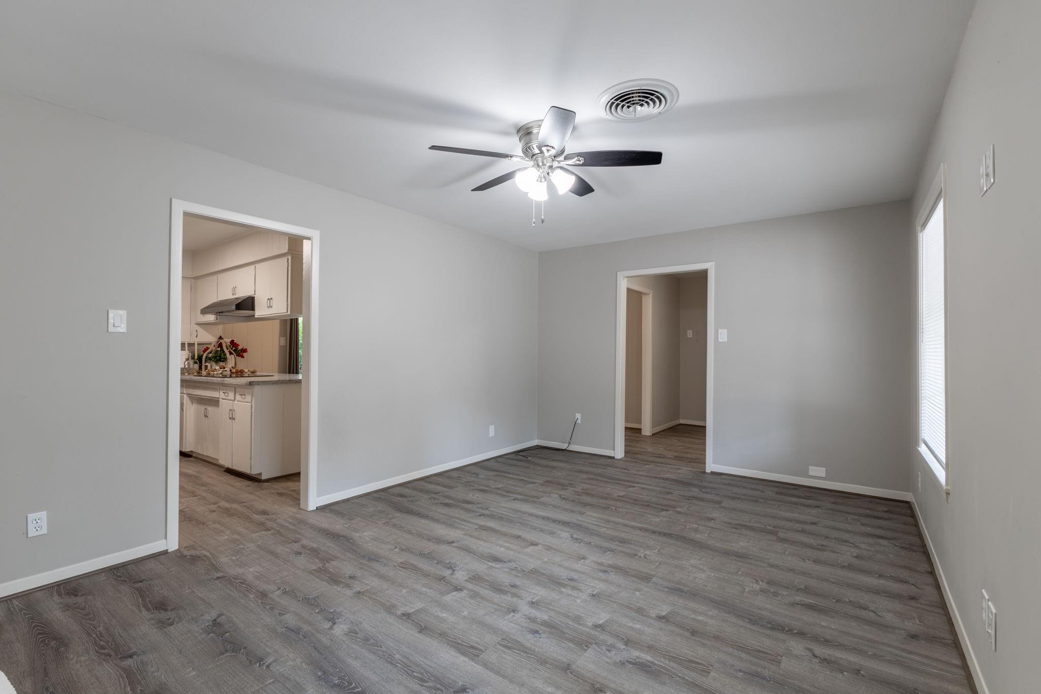 Vacant living room with gray plank flooring, a ceiling fan, and an open doorway to the kitchen.