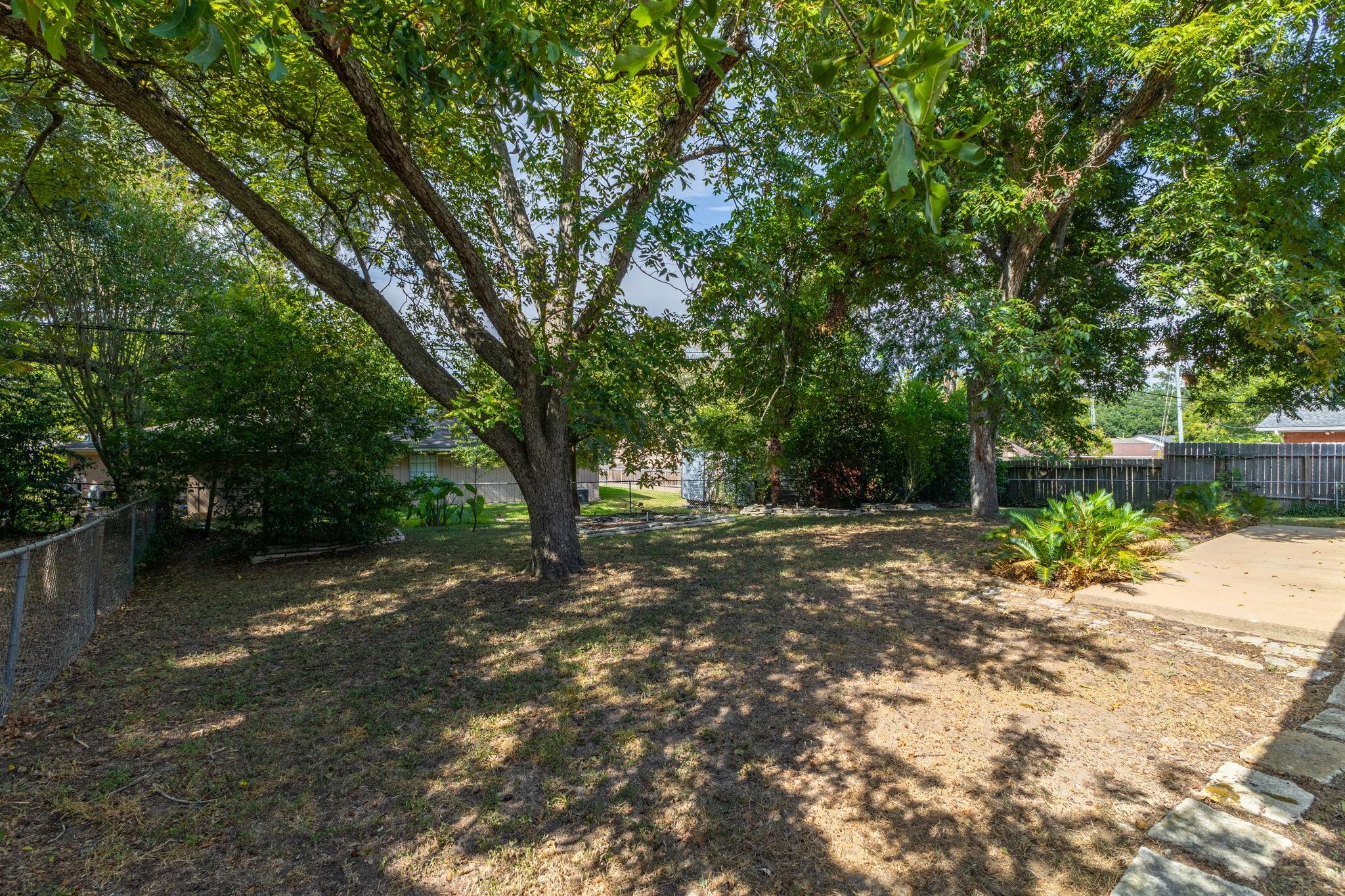 Shaded backyard with mature trees, a fenced lawn, and a concrete patio.