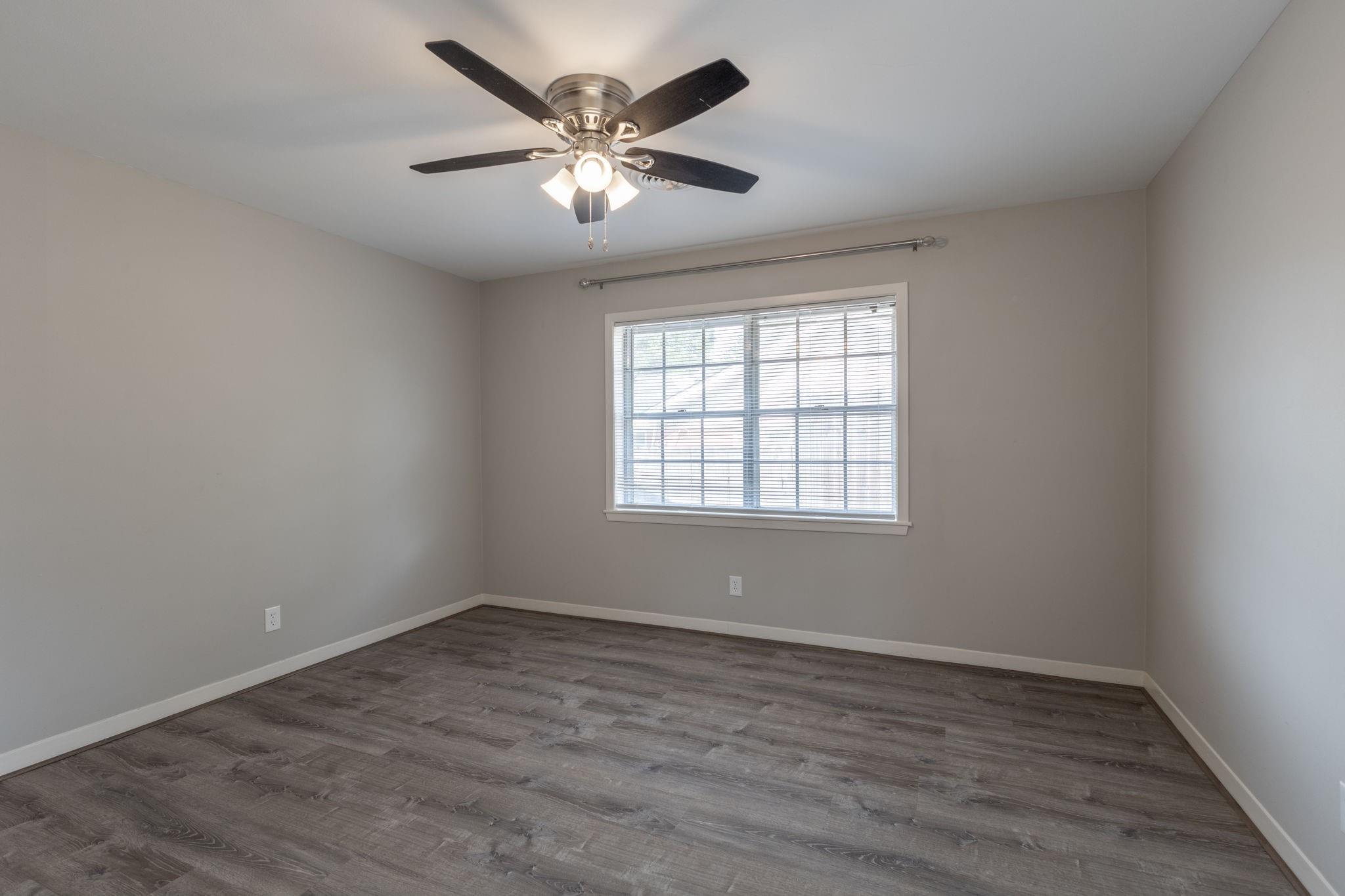 Empty room with gray wood-look floors, a window with blinds, and a ceiling fan with lights.