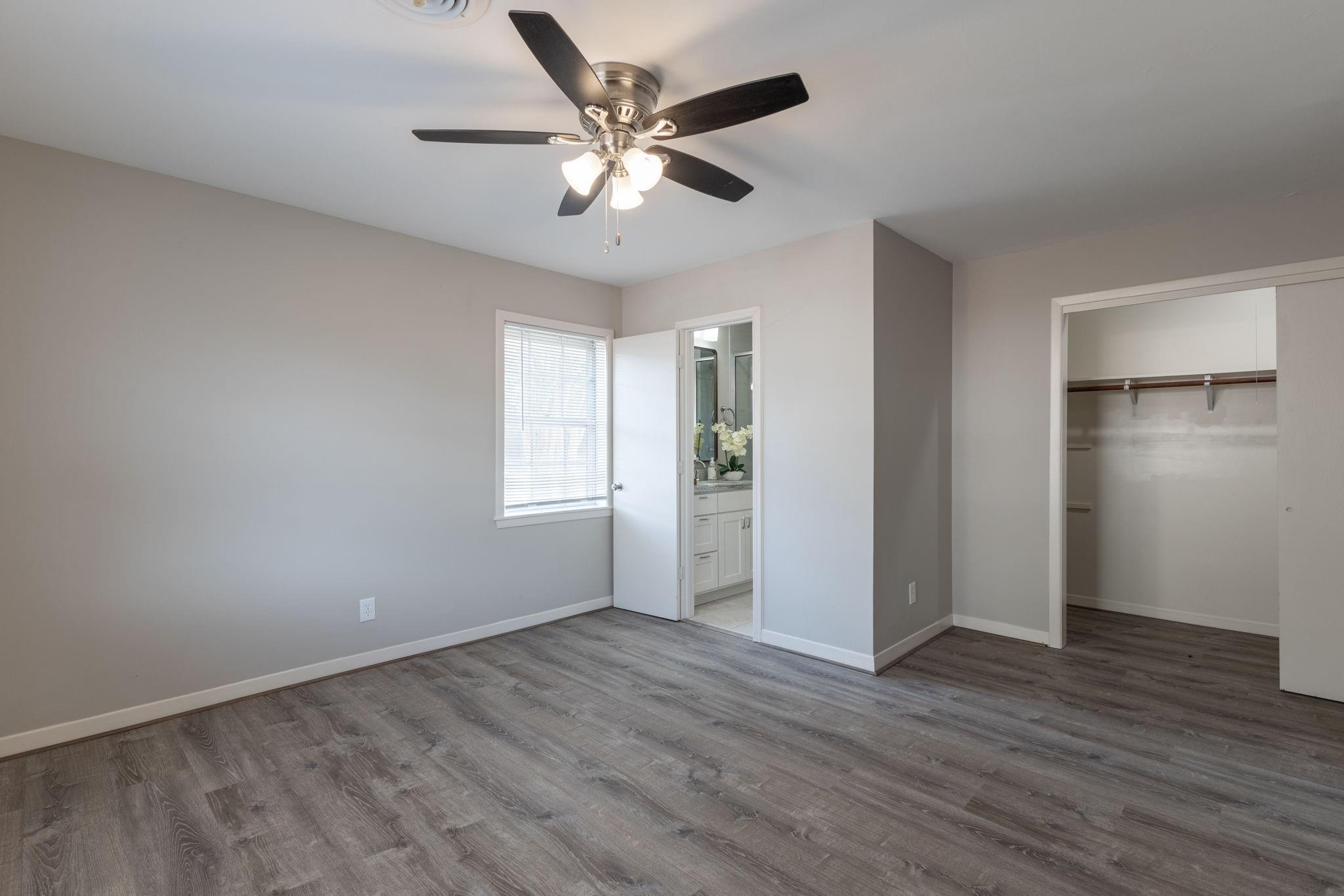 Vacant bedroom with gray flooring, a ceiling fan, an open closet, and a bathroom doorway.