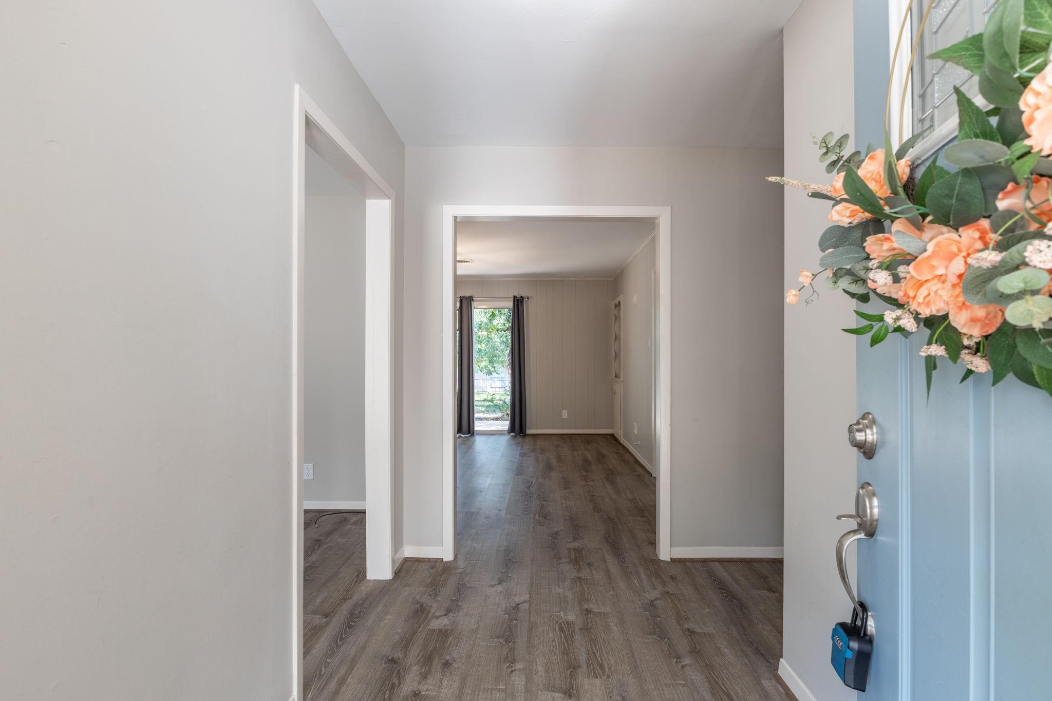 Entryway with dark plank floors leading to a vacant room and glass door; light-blue front door has a floral arrangement.
