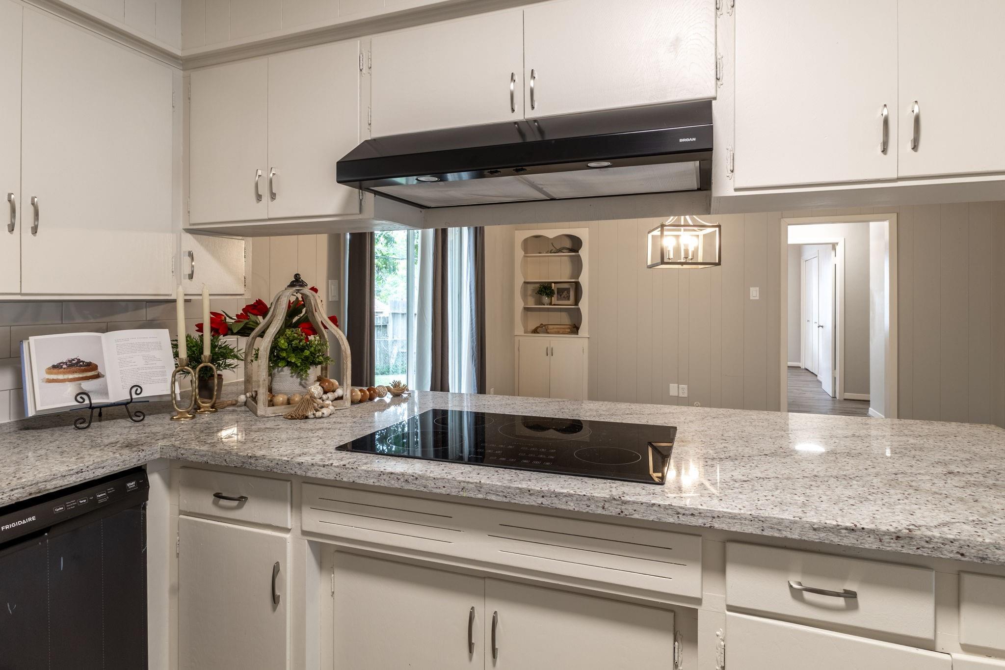 Kitchen with white cabinets, granite counters, a black cooktop, and red flowers on the counter.