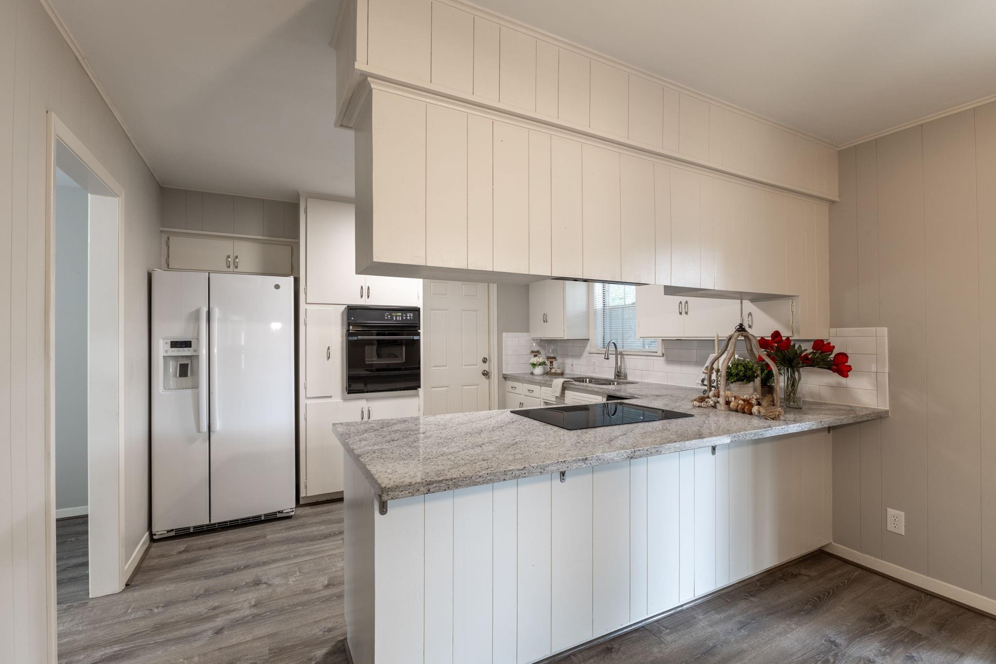 Kitchen with white cabinets, granite island and cooktop, stainless refrigerator, and gray wood-look floors.