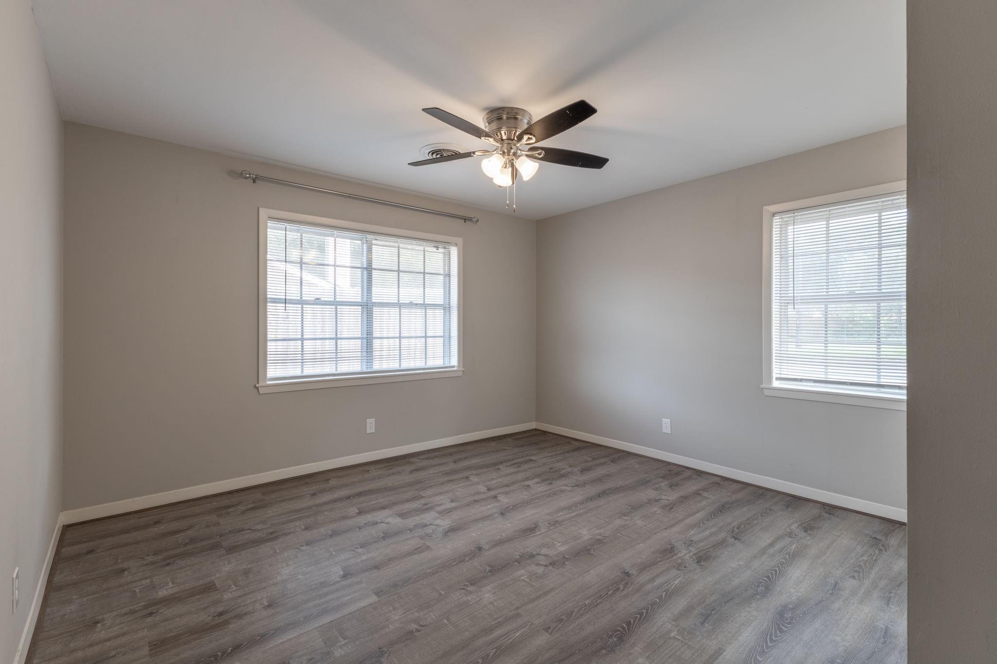 Empty room with gray wood-look flooring, two windows with blinds, and a ceiling fan.