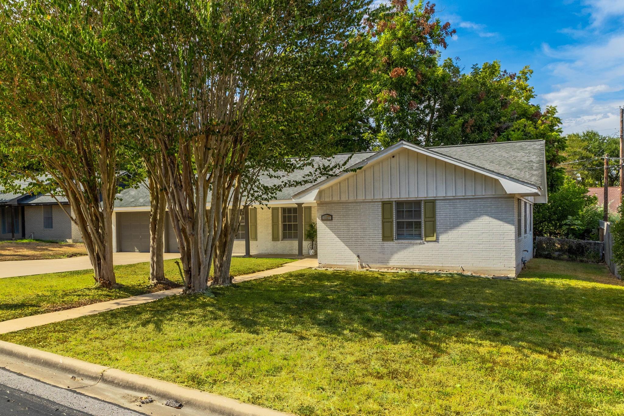 Single-story brick house with green shutters, attached garage, and mature trees shading the front lawn.