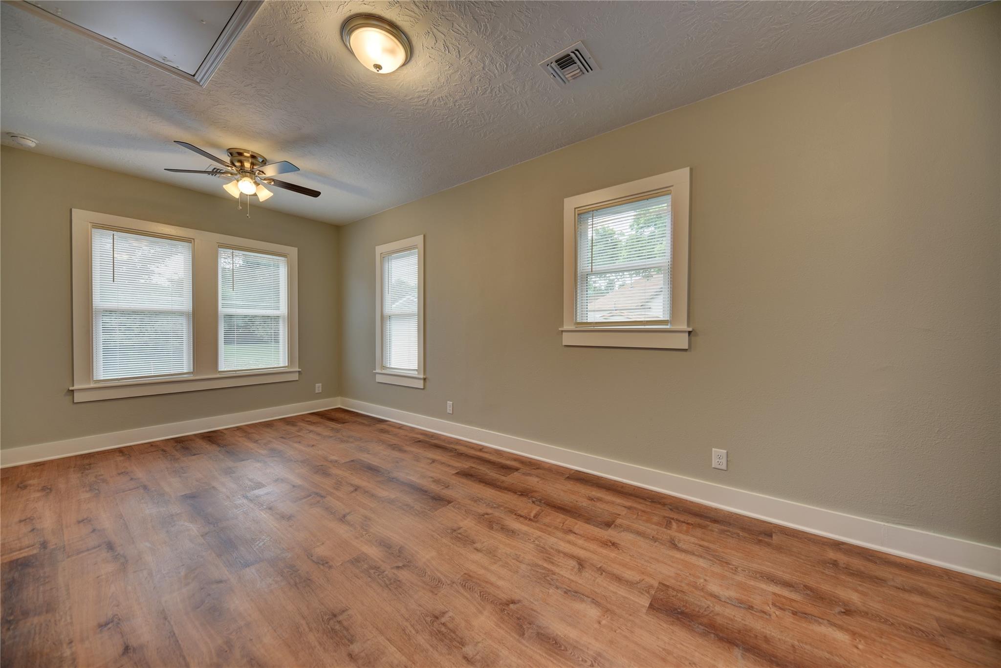 Empty room with wood-look floors, beige walls, four windows, and a ceiling fan.