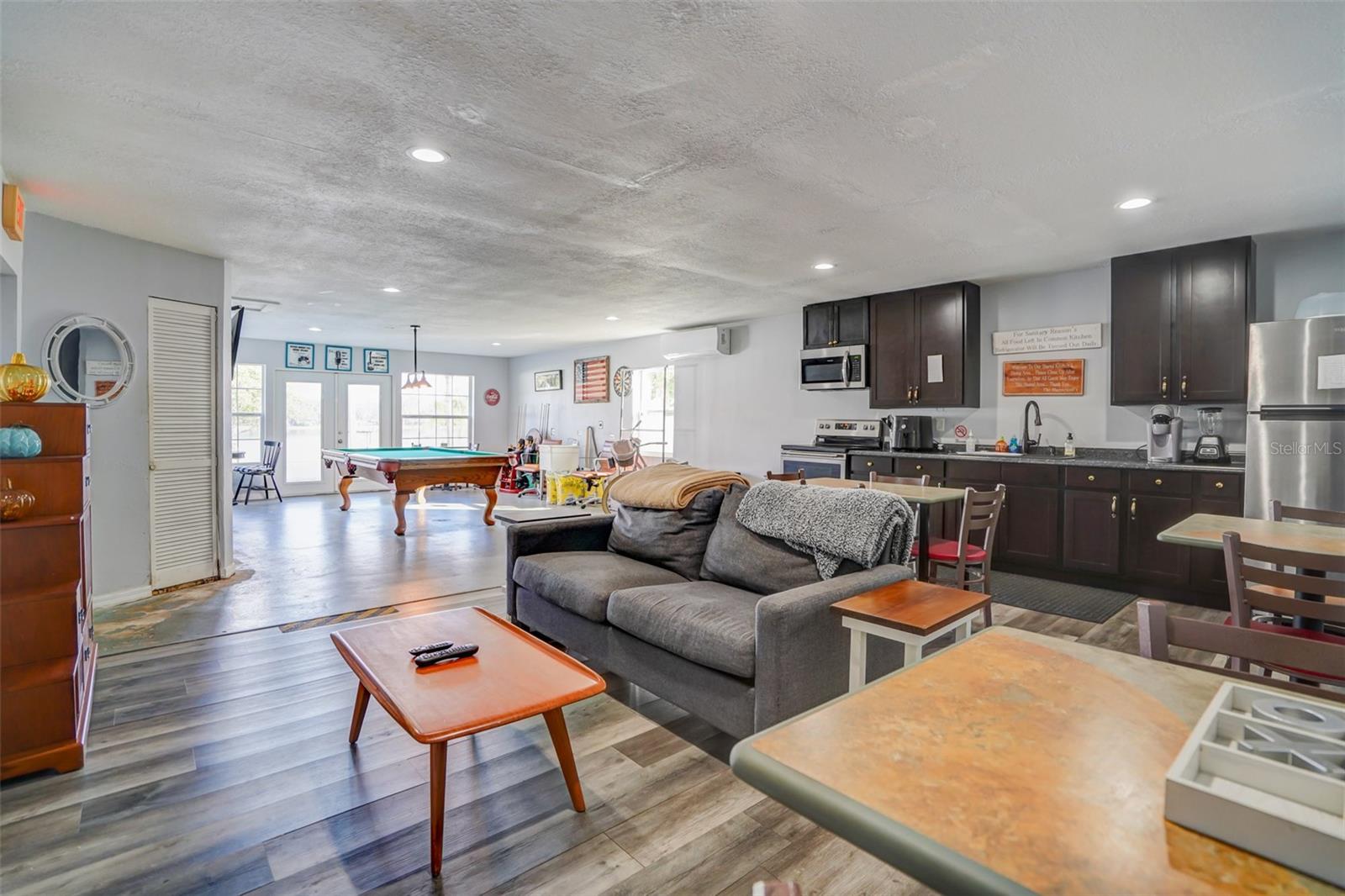 Open living and kitchen area with dark cabinets, gray sofa, and a pool table by the glass doors.
