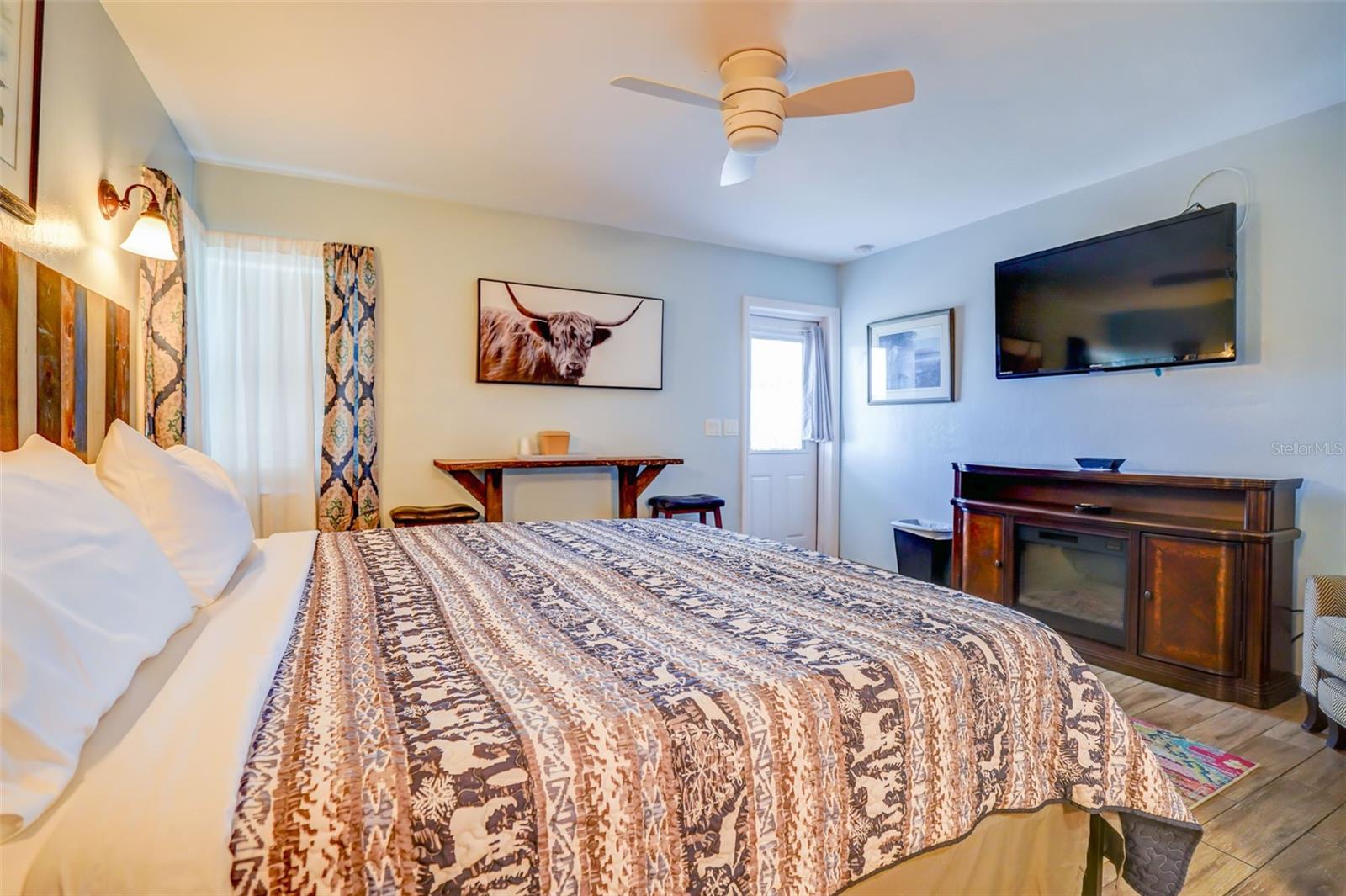 Furnished bedroom with a patterned bedspread, ceiling fan, wall-mounted television, and dark wood dresser.