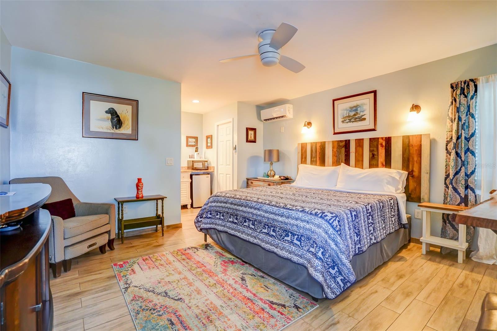 Furnished bedroom with patterned bedding, a multicolored wood headboard, and a compact kitchenette.