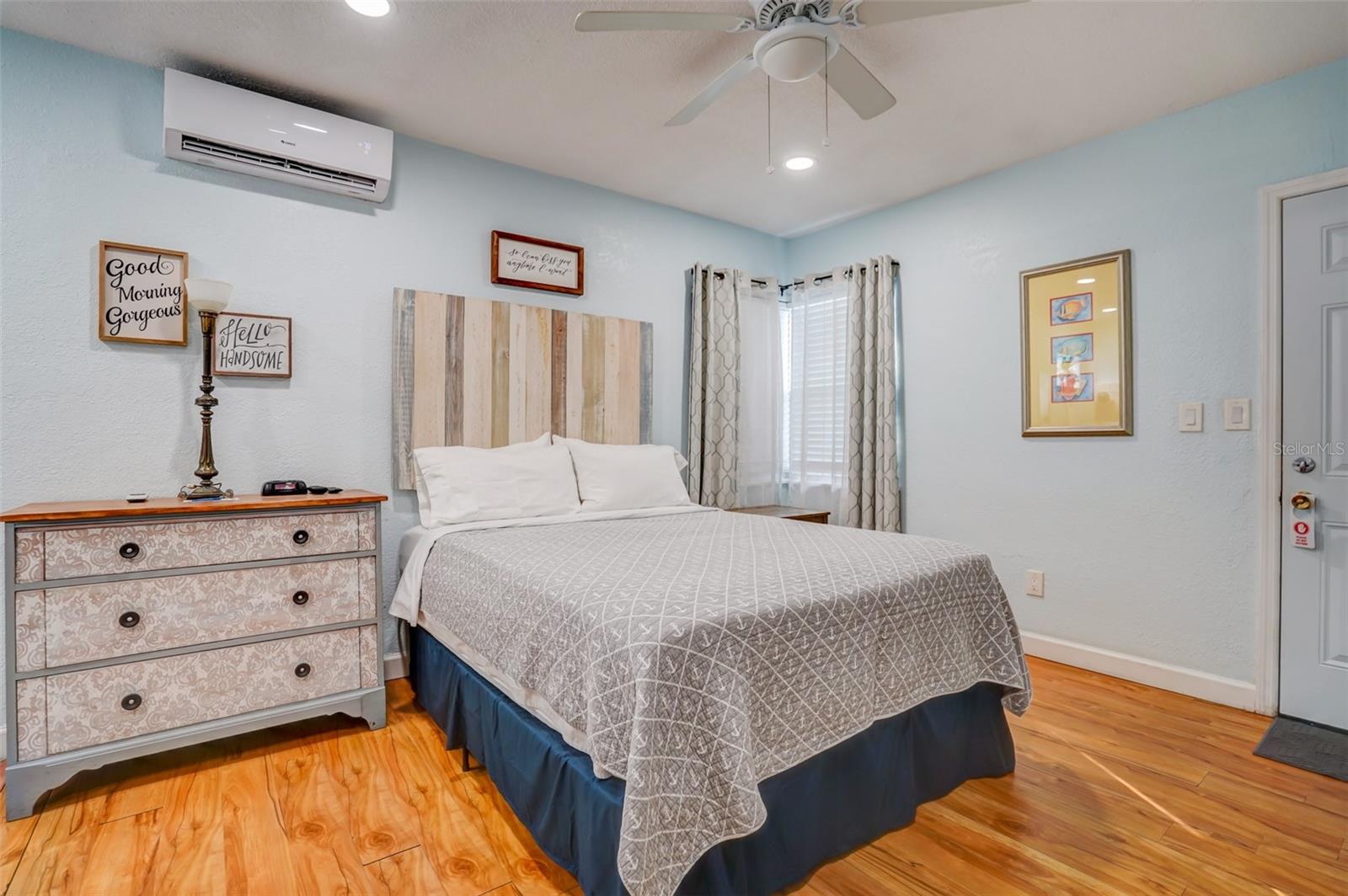 Bedroom with a gray quilted bed, wood headboard, patterned dresser, and warm wood flooring.