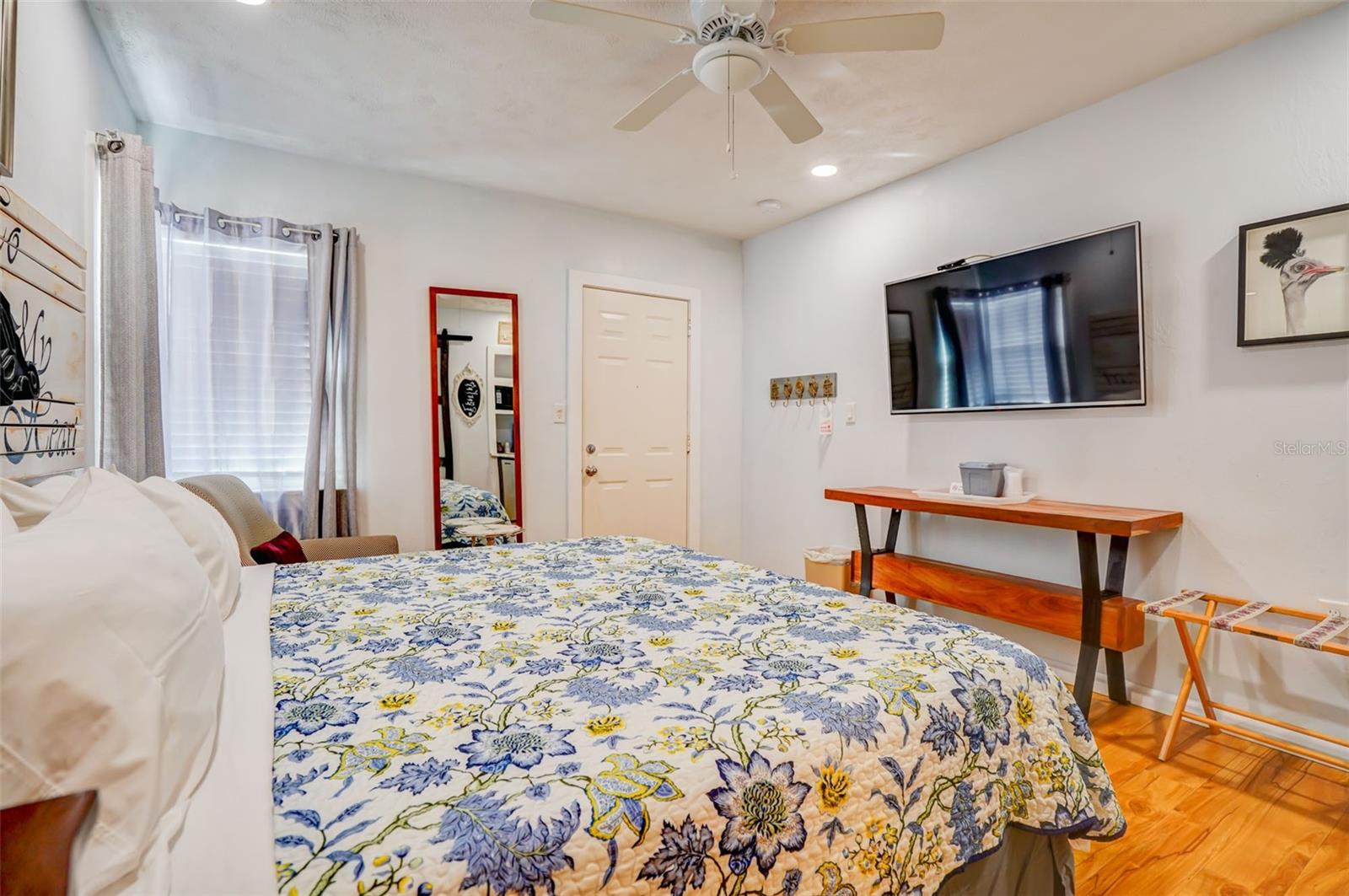 Furnished bedroom with a floral bedspread, wood flooring, ceiling fan, and wall-mounted television.