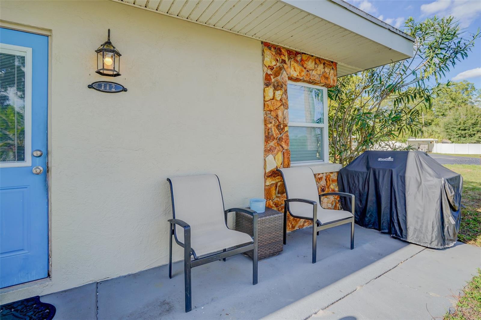 Covered porch with a blue door, stone-trimmed window, patio chairs, and covered grill.