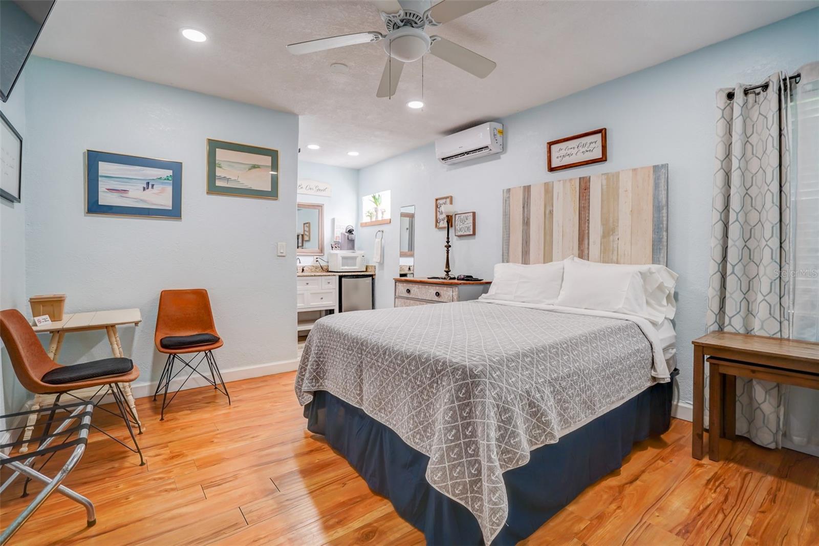 Bedroom with a large gray-quilted bed, pale blue walls, wood-look flooring, and an adjoining vanity.