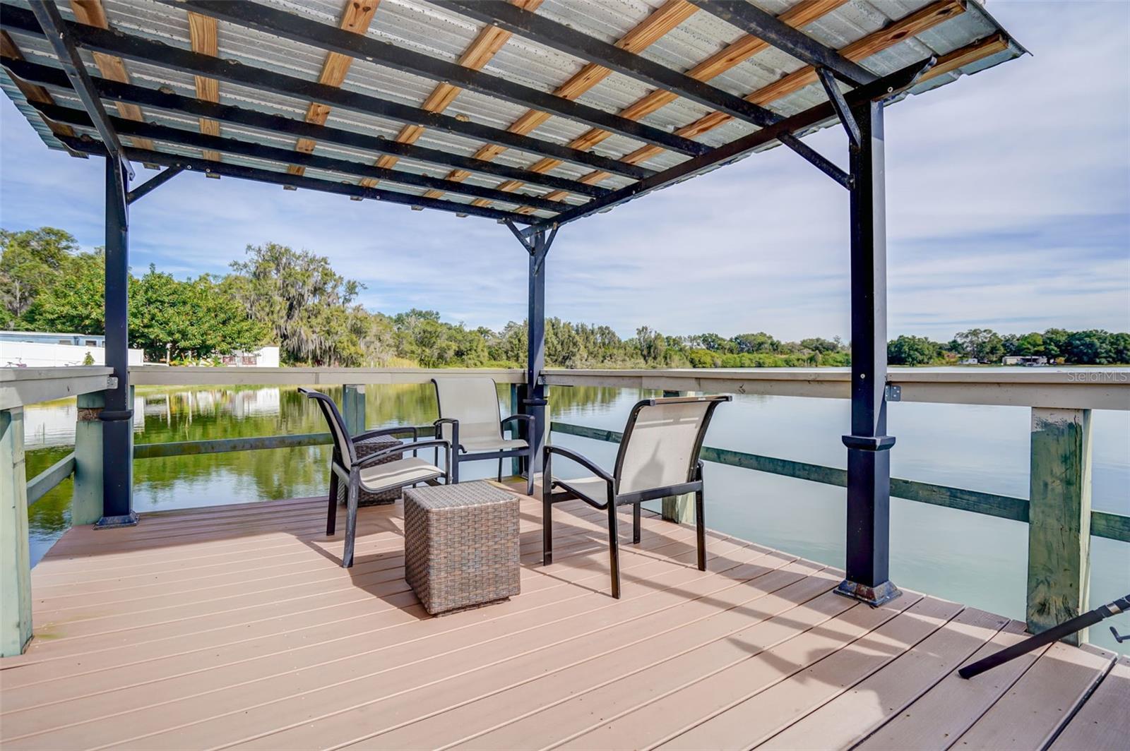 Covered waterfront deck with patio seating overlooking calm water and a tree-lined shore.