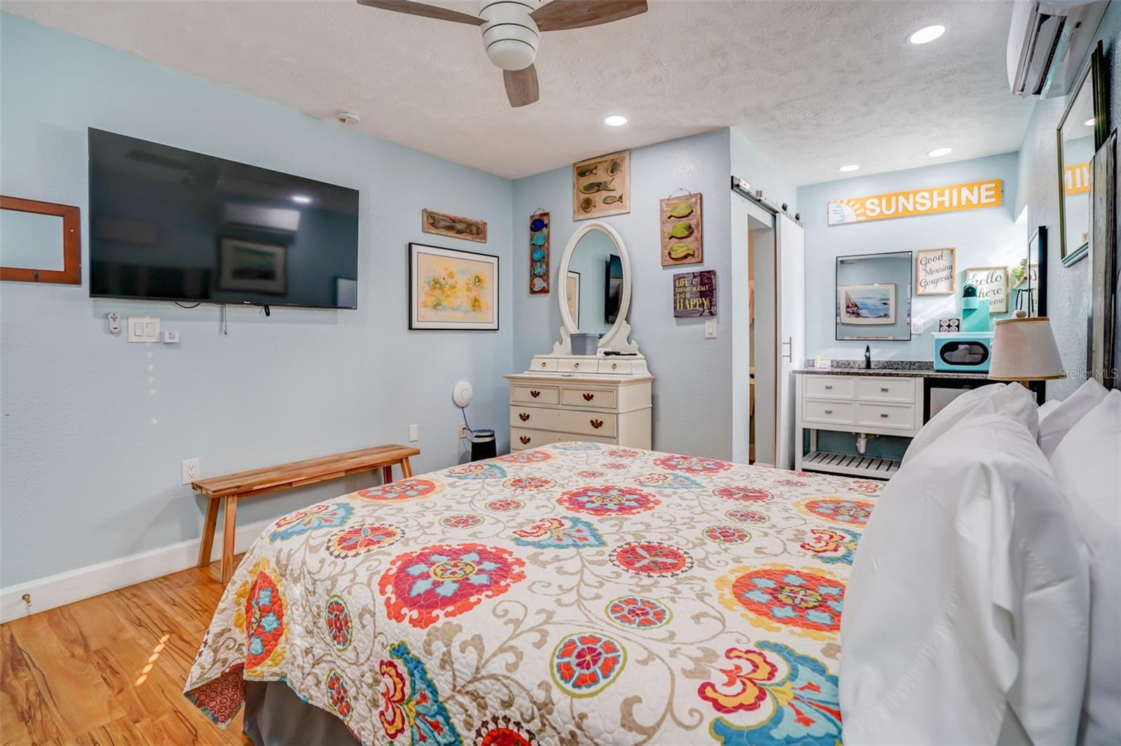 Furnished bedroom with a colorful floral bedspread, wall-mounted television, and white dresser with oval mirror.