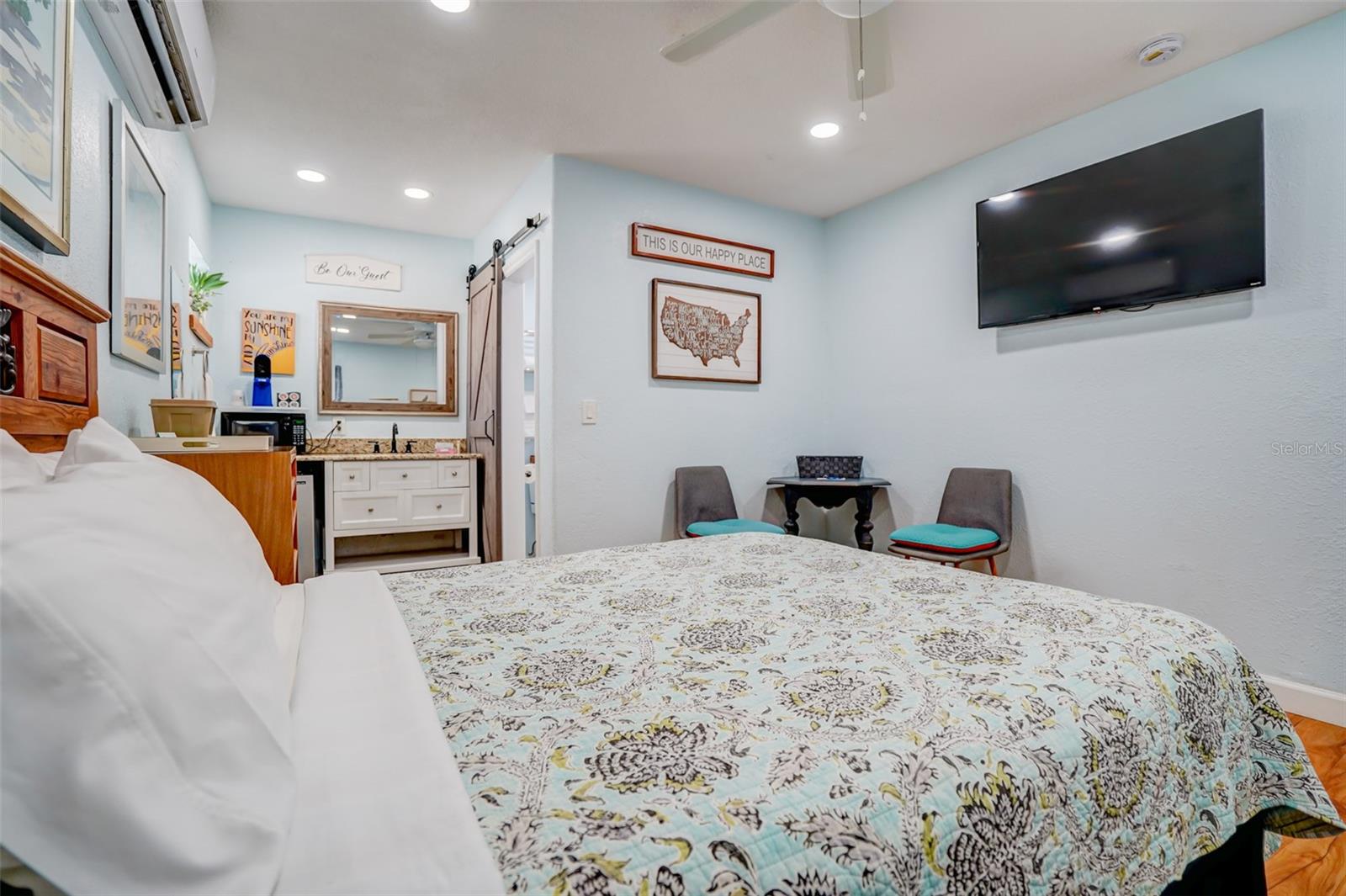 Furnished bedroom with a patterned bedspread, wall-mounted TV, two chairs, and an adjoining vanity area.