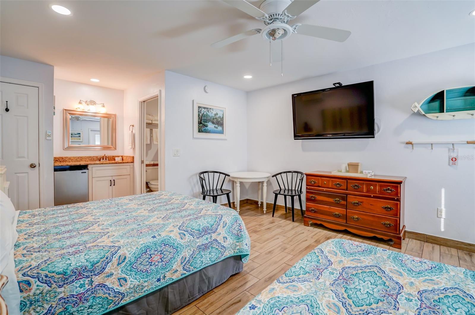 Bedroom with two patterned beds, a wall-mounted television, and a vanity beside the bathroom doorway.