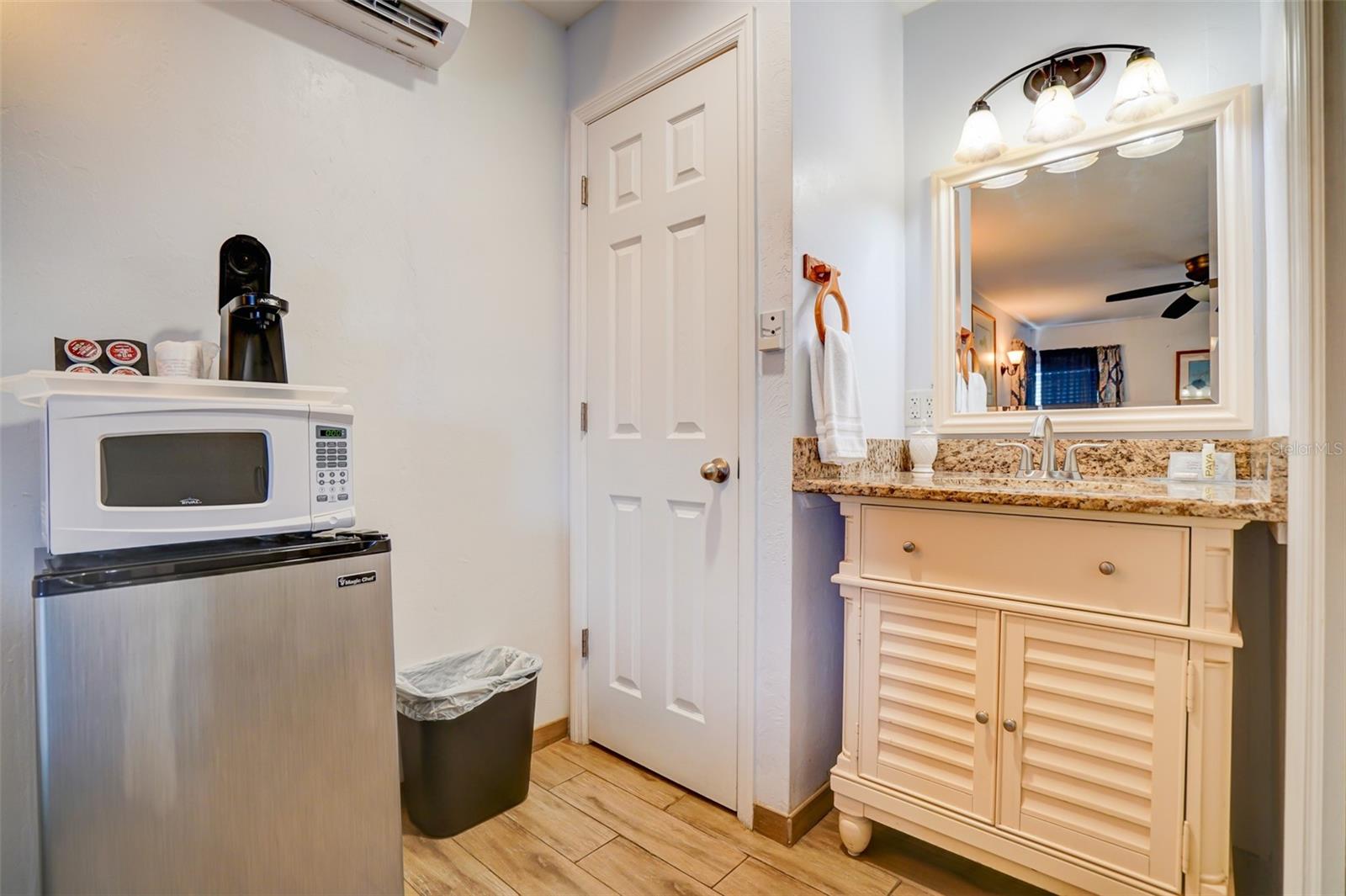 Bathroom vanity with granite counter and mirror beside a microwave and compact refrigerator.