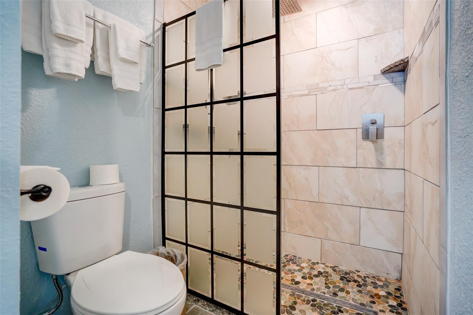 Bathroom with a black grid-glass shower, pale tiled walls, pebble floor, and white toilet.