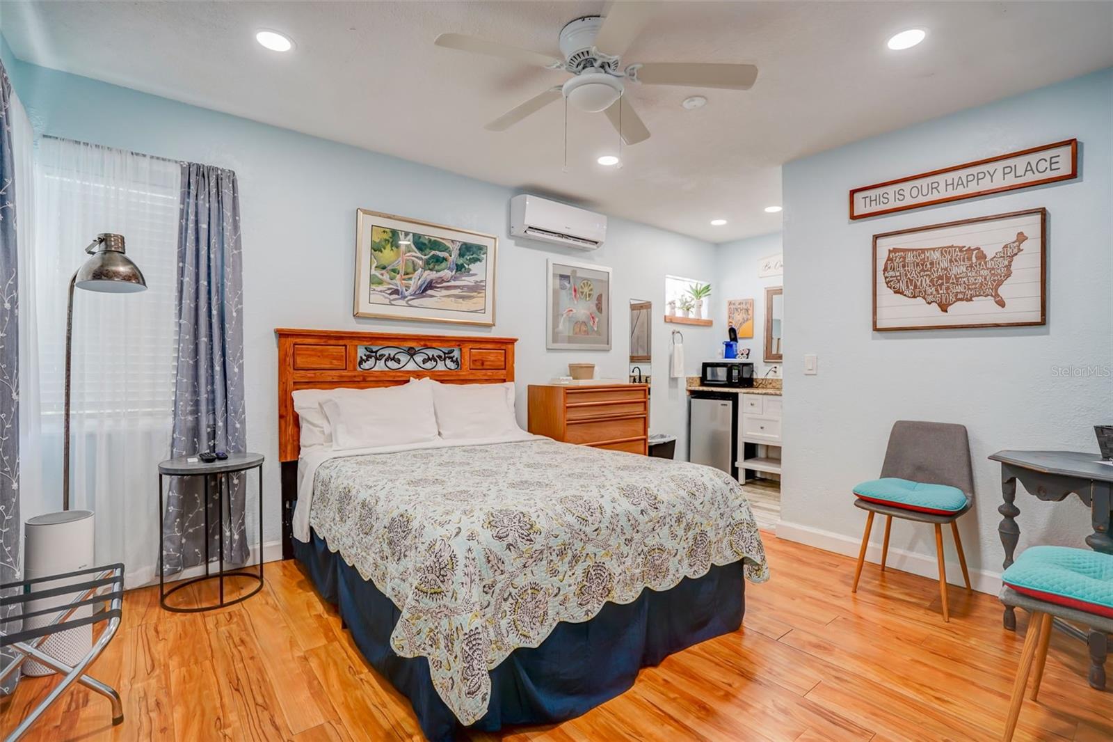 Furnished bedroom with a large bed, wood flooring, ceiling fan, and small kitchenette area.