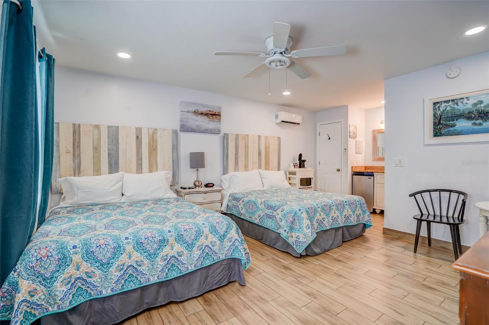 Bedroom with two beds, patterned blue bedding, teal curtains, and wood-look tile flooring.