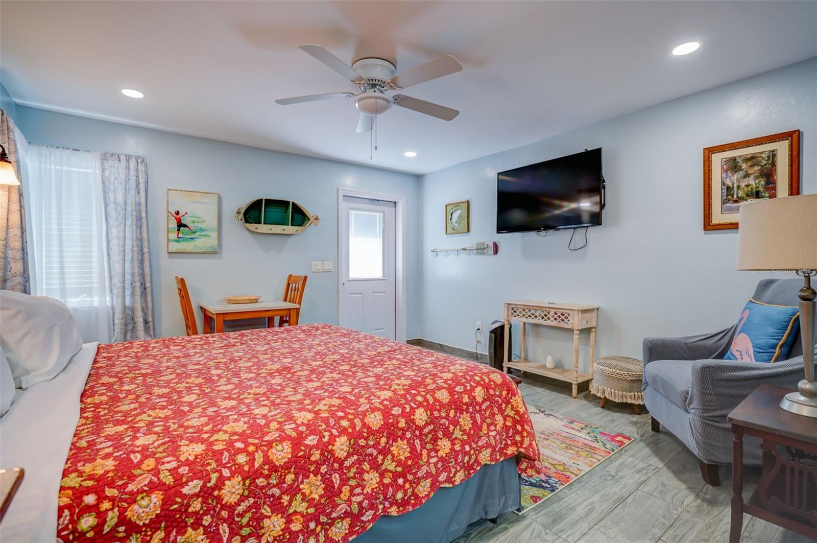 Bedroom with a red floral bedspread, wall-mounted television, ceiling fan, and blue armchair.