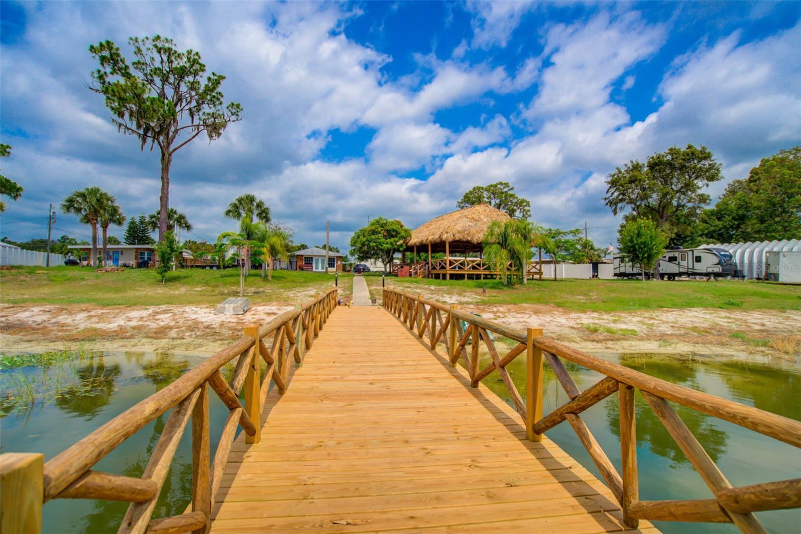 Wooden boardwalk crosses a pond toward a thatched-roof pavilion, with palm trees and RVs beyond.