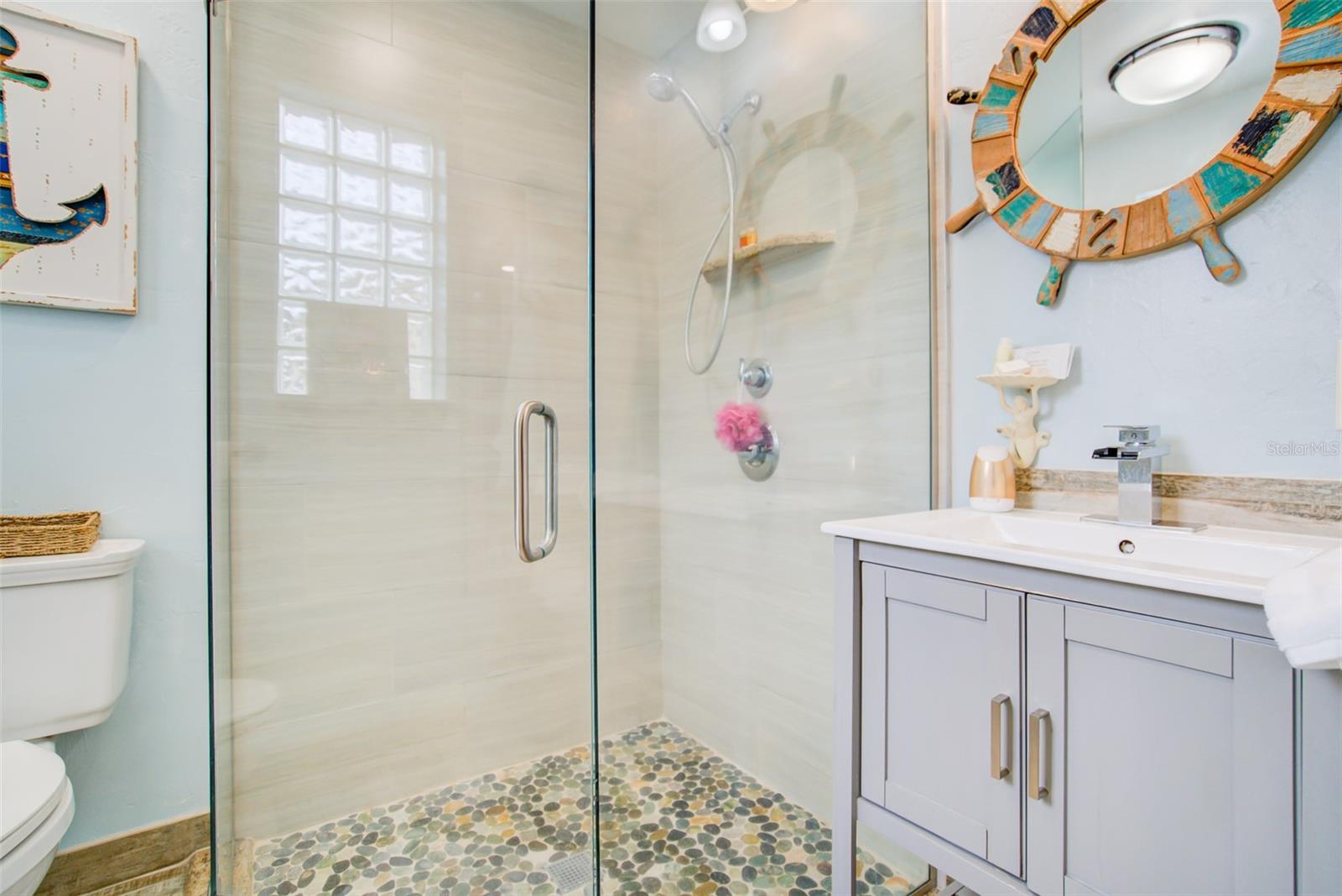 Bathroom with a glass shower, pebble-stone floor, and gray vanity beneath a round mirror.