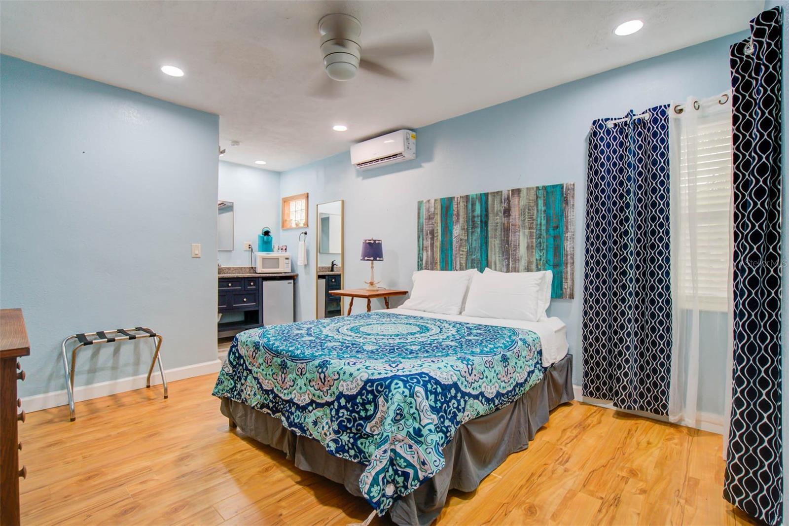 Furnished bedroom with a blue patterned bedspread, wood-look floors, and a kitchenette nook.