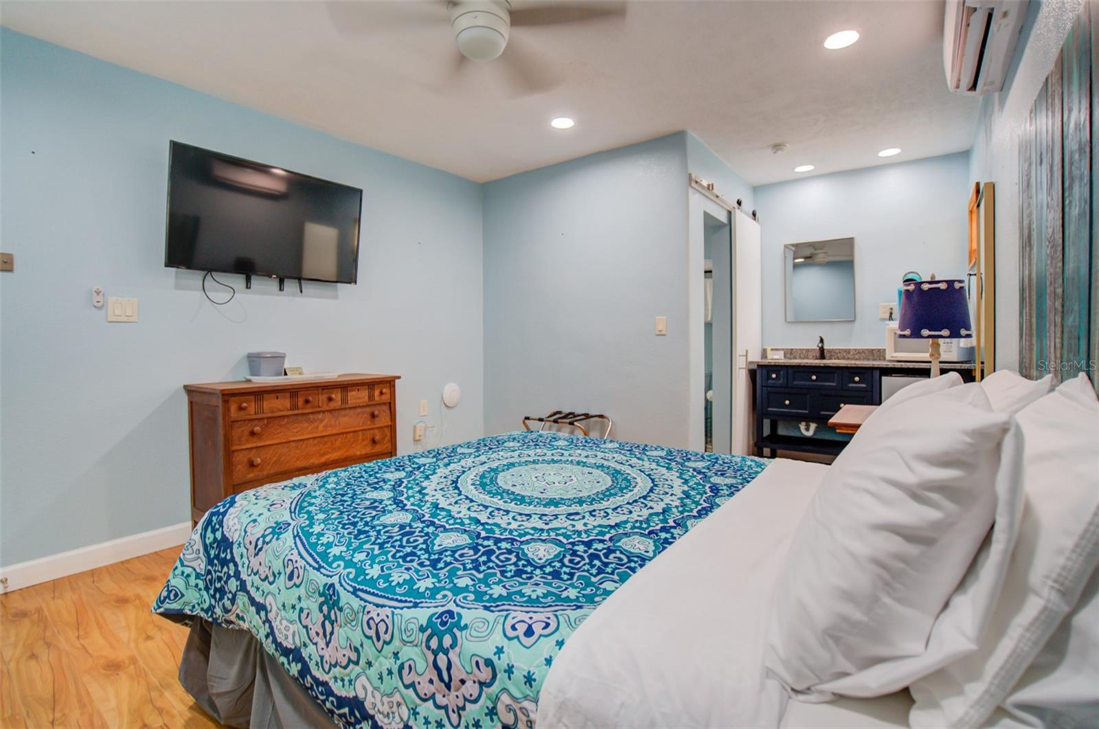 Furnished bedroom with a blue patterned bedspread, wall-mounted TV, wooden dresser, and adjoining vanity.