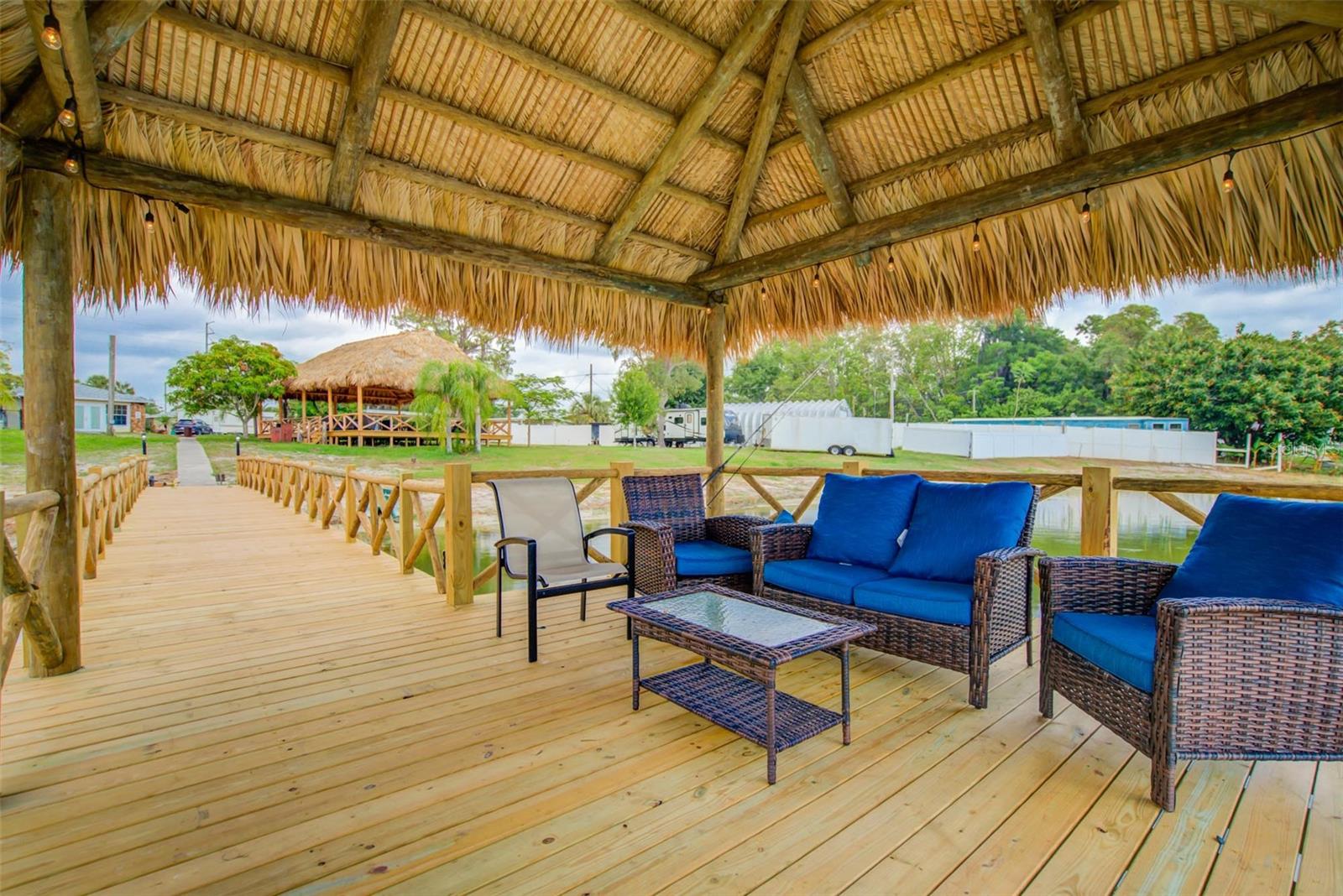 Thatched-roof deck with blue-cushioned wicker seating beside a wooden walkway and water.